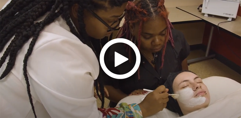 Video play button with an esthetician instructor guiding a student on how to apply a skincare product to the face. 
