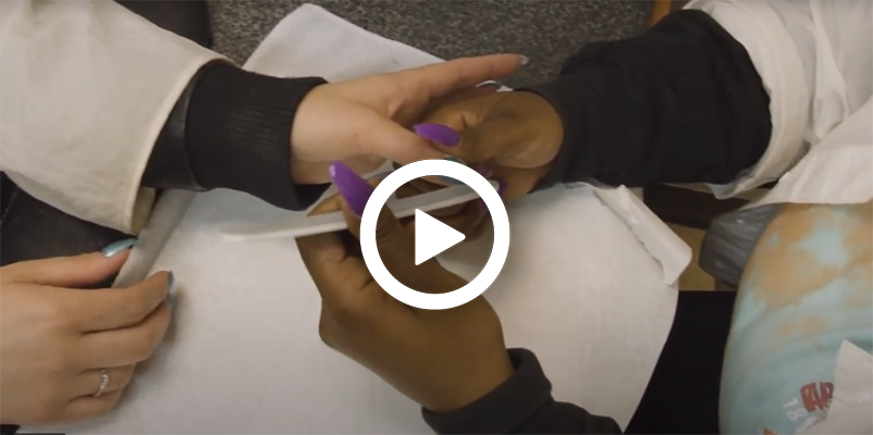 Video play button with a manicuring professional teaching a student how to properly work on nails. 